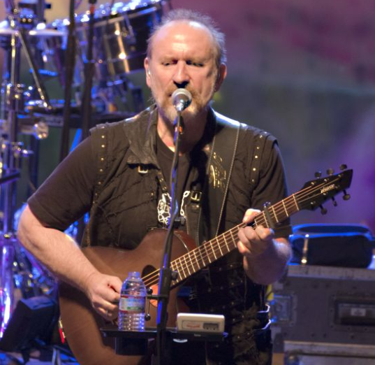 Musician Colin Hay singing and playing acoustic guitar.