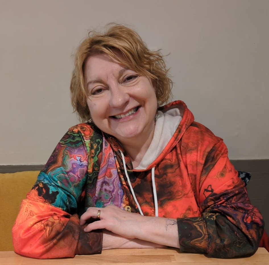 My wife Gina sitting at a table, wearing a colorful hoodie and smiling.