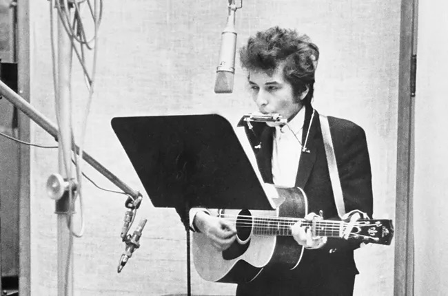 Bob Dylan with guitar and harmonica in the recording studio, standing in front of a music stand.