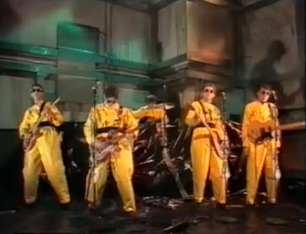 Group shot of the band Devo on stage with yellow hazmat like suits