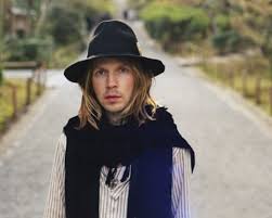 Singer Beck outdoors standing in the middle of a countryish looking road, wearing black scarf and hat.
