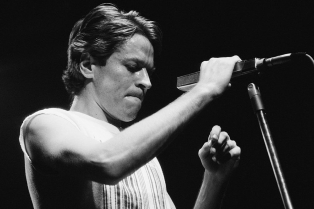 Singer Robert Palmer in a sleeveless tea holding a microphone on a stand.