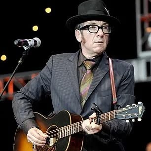 Elvis Costello on stage with suit and tie and hat playing acoustic guitar