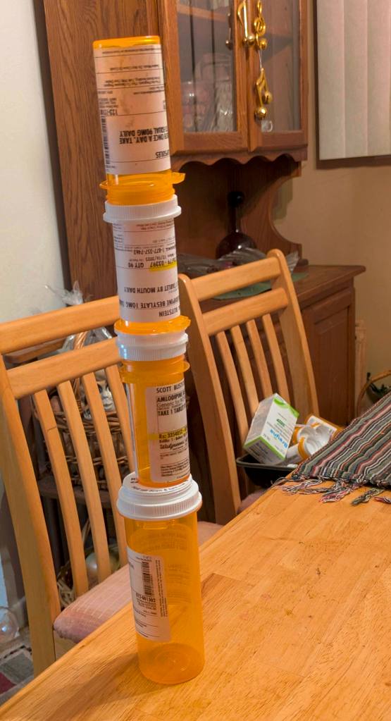 4 empty prescription pill bottles stacked one of top of another on our dining room table - just daring one of our cats to know it down.