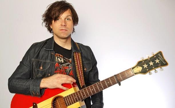 Ryan Adams holding red and yellow striped acoustic guitar in front of white background.