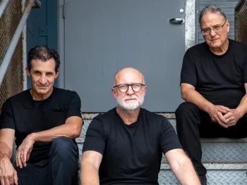 Photo of the band Sugar seated on steps in front of gray door. Photo by Ryan Bakerink