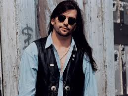 Steve Earle outdoors in blue shirt and black vest - sunglasses.