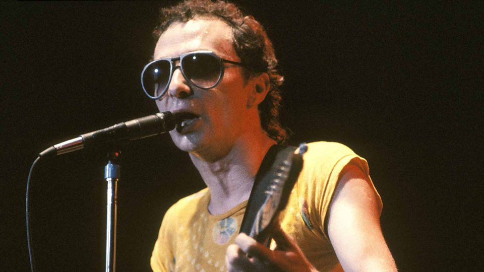 Graham Parker at microphone holding guitar and singing, wearing sleeveless tee and dark sunglasses.