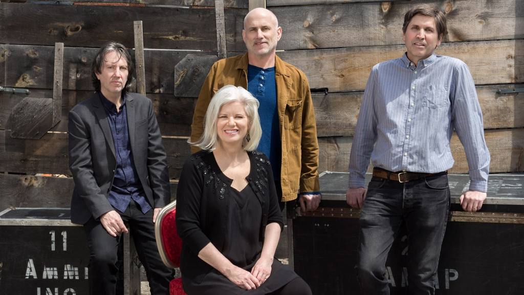 Group shot of the band Cowboy Junkies seated outdoors in front of brick wall.