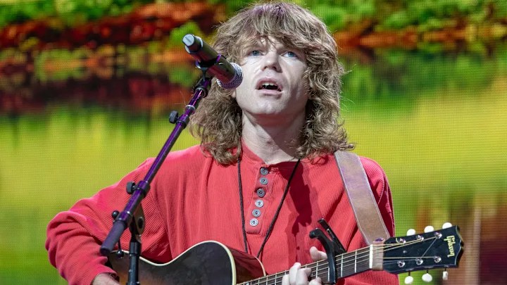Singer Jesse Welles in front of microphone, holding acoustic guitar. Wearing a red shirt in front of outdoors backdrop.