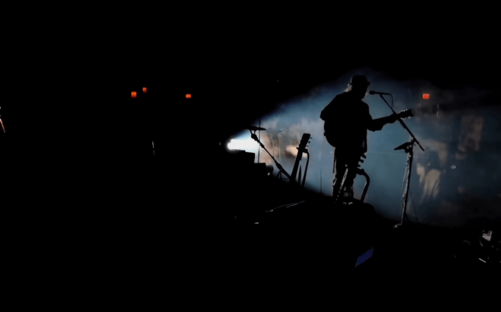 Neil Young on a dark stage with very slight lighting focused on him from behind,