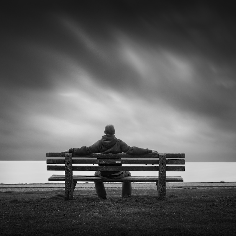 man sitting on bench with his arms stretched outward on the back of the bench, they sky is dark and ominous looking.